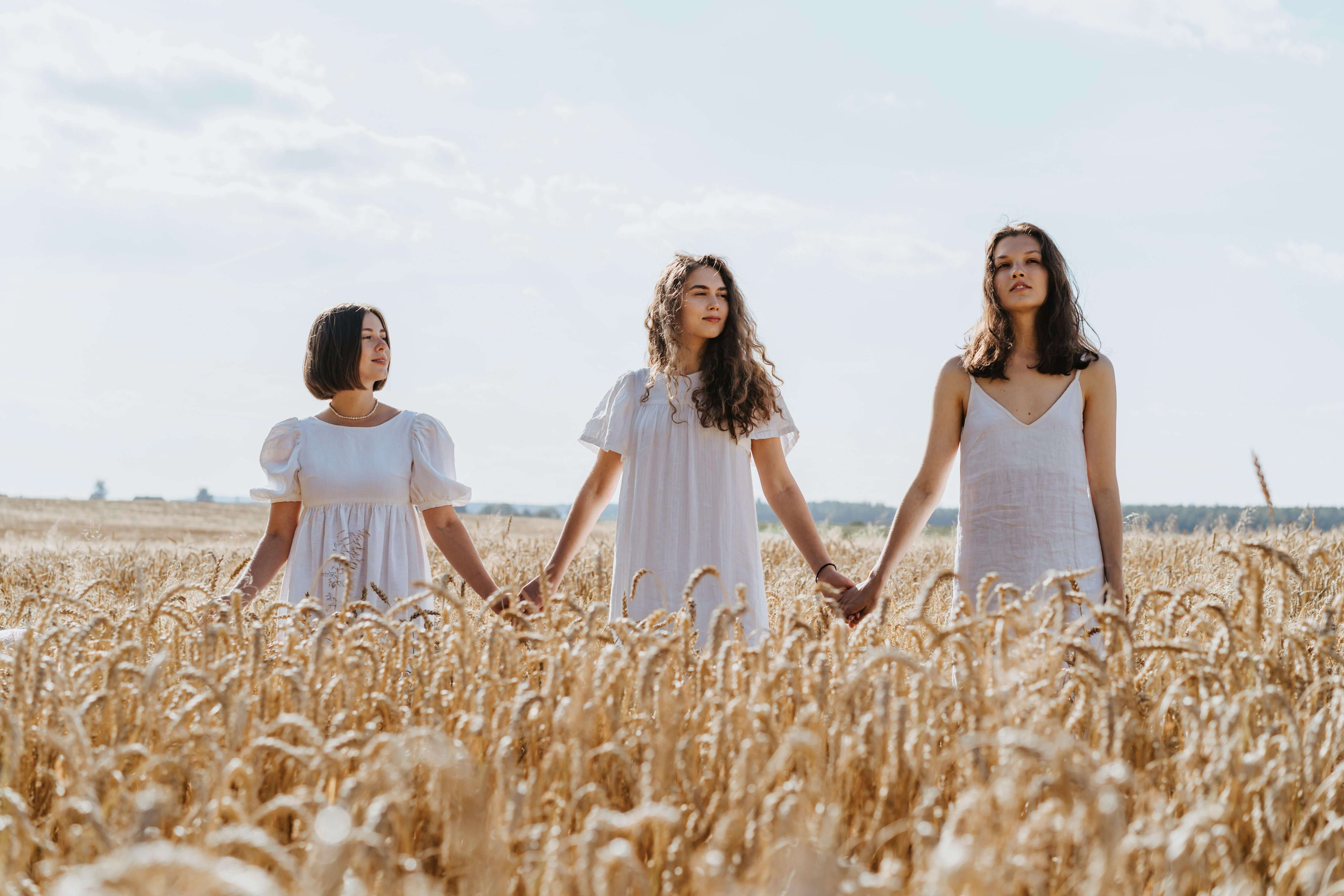 Three women in white dresses holding hands in a golden wheat field under blue sky
